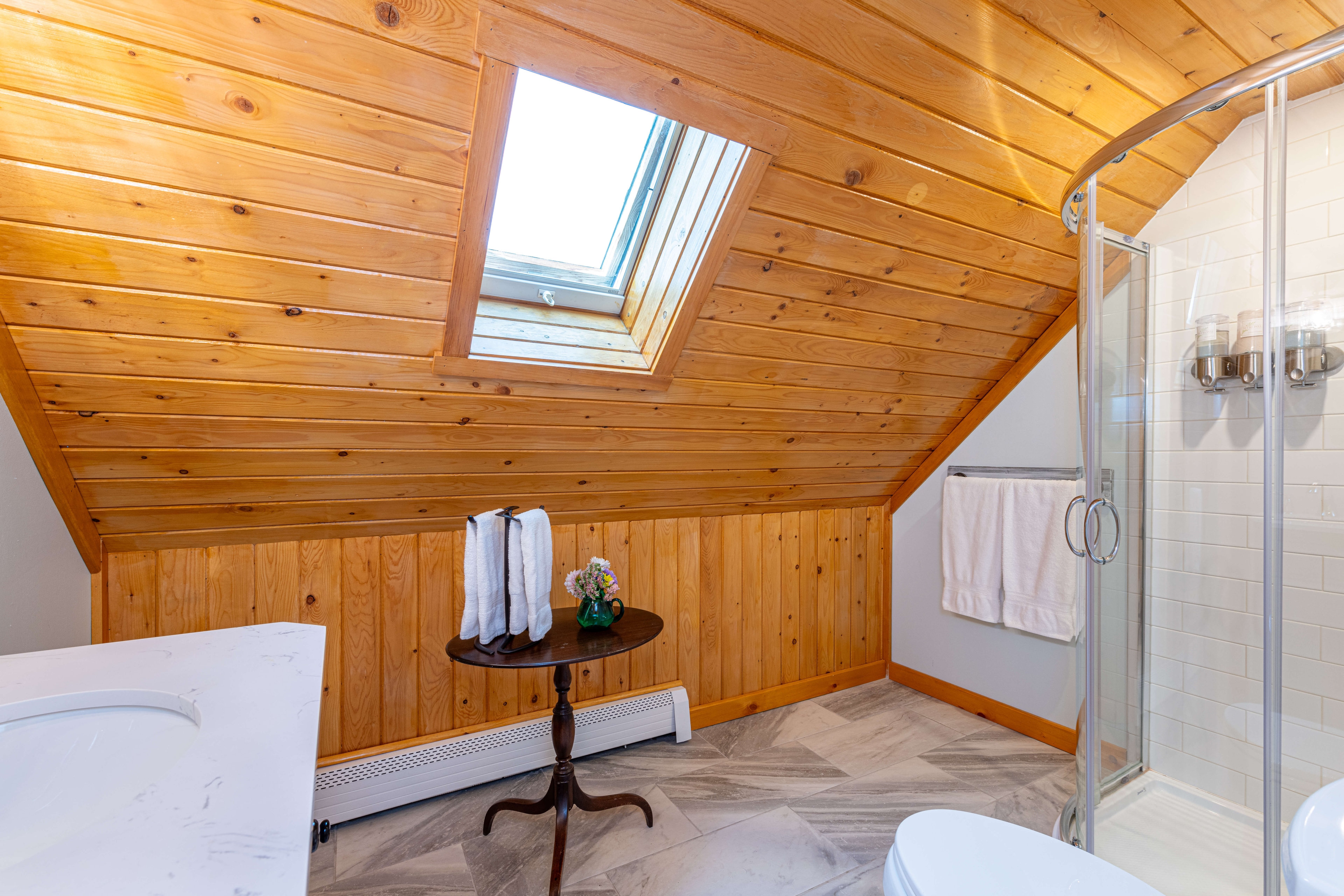 Glass enclosed shower with gray tile, towel rack with plush white towels, slanted wood ceiling with skylight