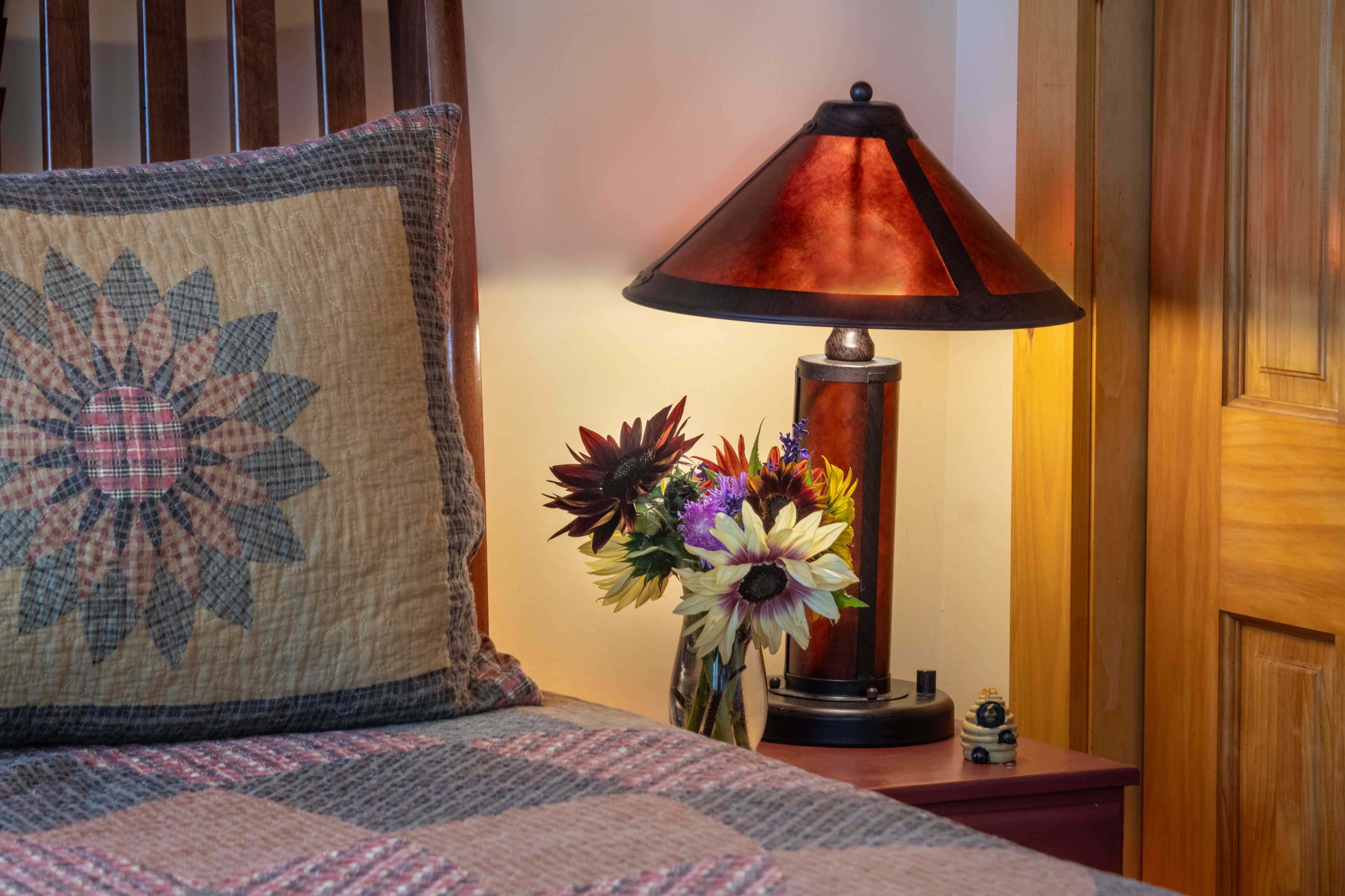 Close up of bed with sunflower pillow next to a nightstand with a lamp and bouquet of flowers.