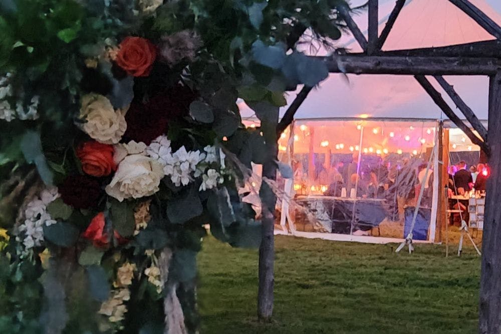 View of an outdoor reception tent full of people at night near a wood arbor full of flowers and greenery