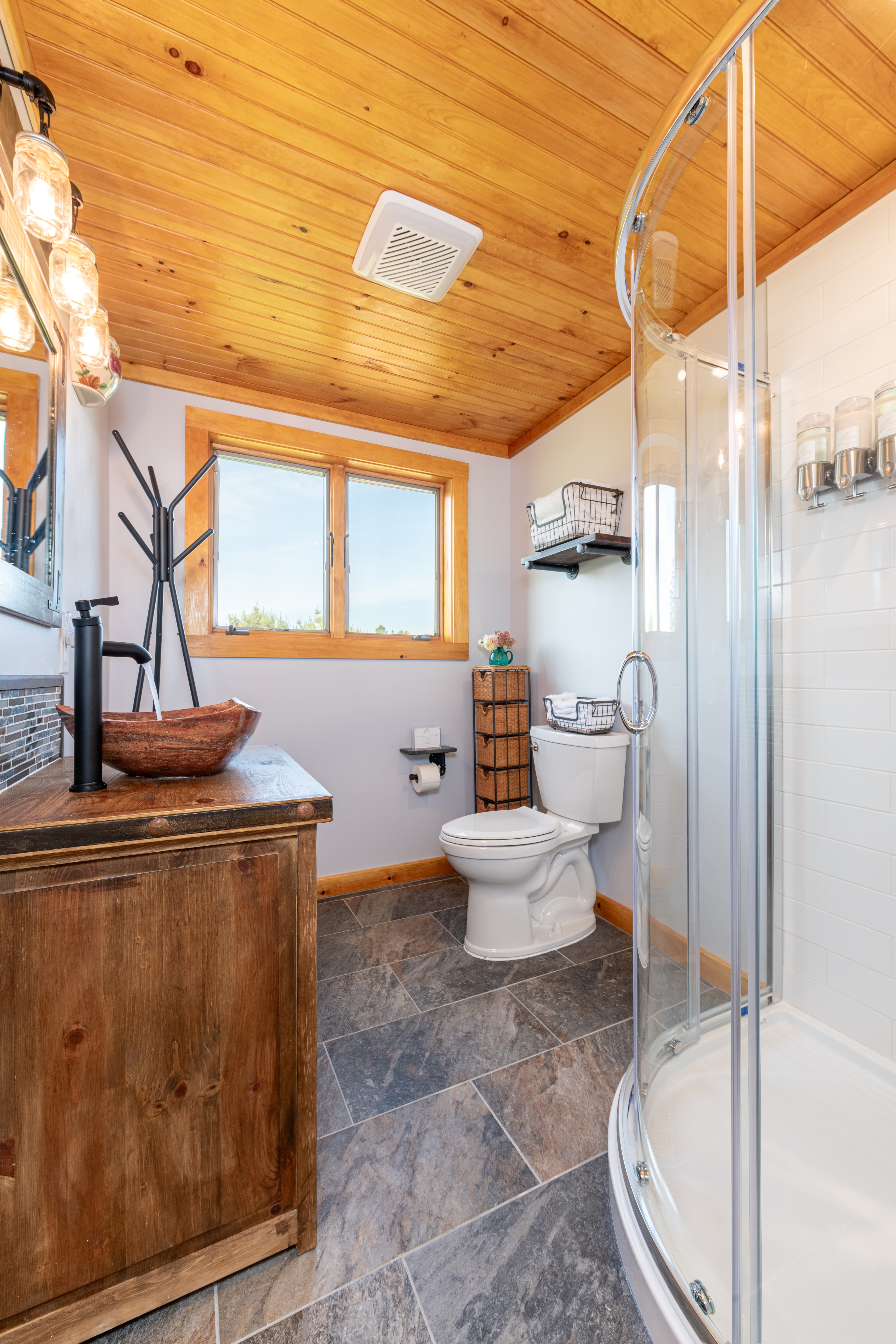 A wooden vanity with an vessel sink, wall mirror, toilet, and glass shower