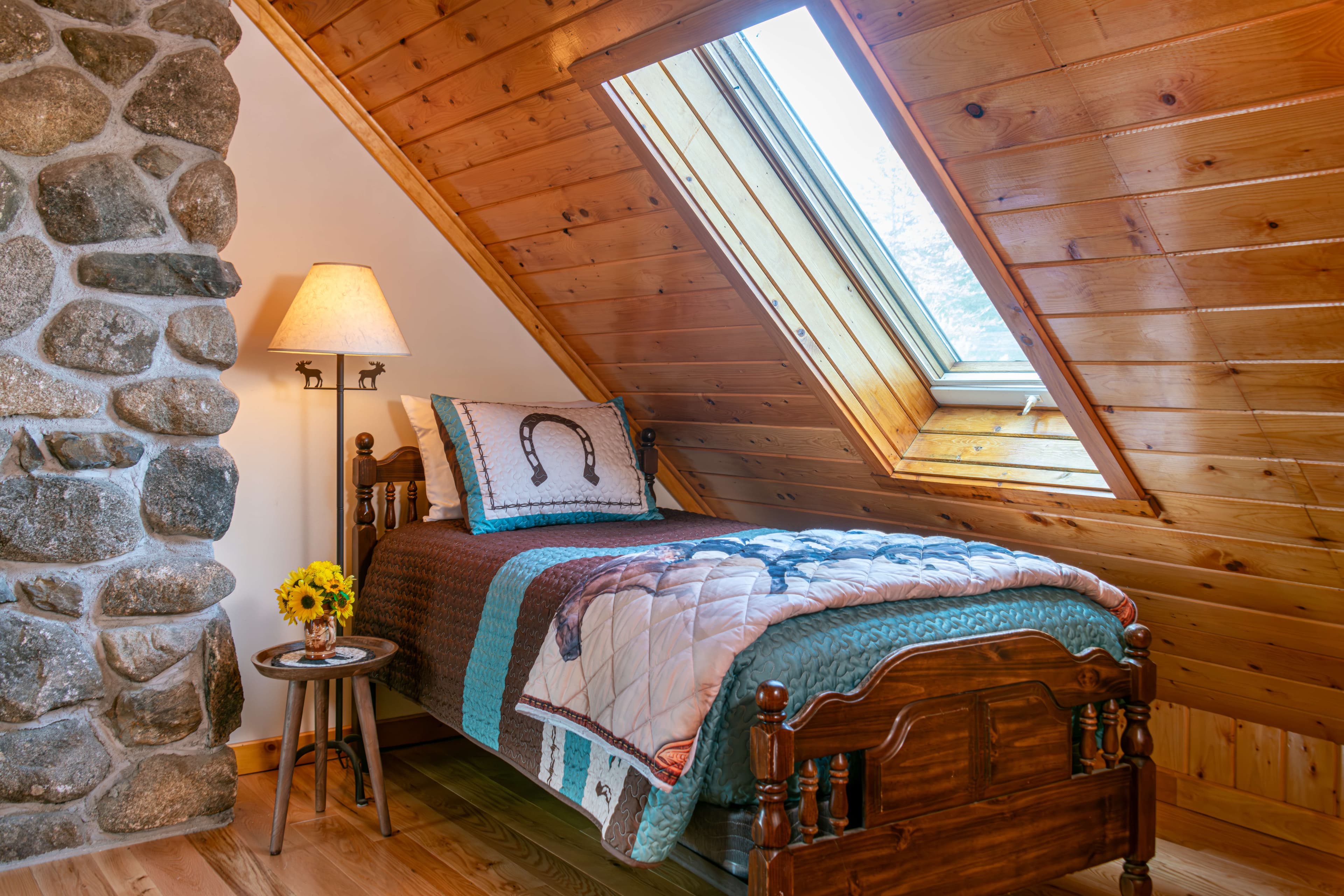 Twin bed under slanted finished wood ceiling next to stone fireplace