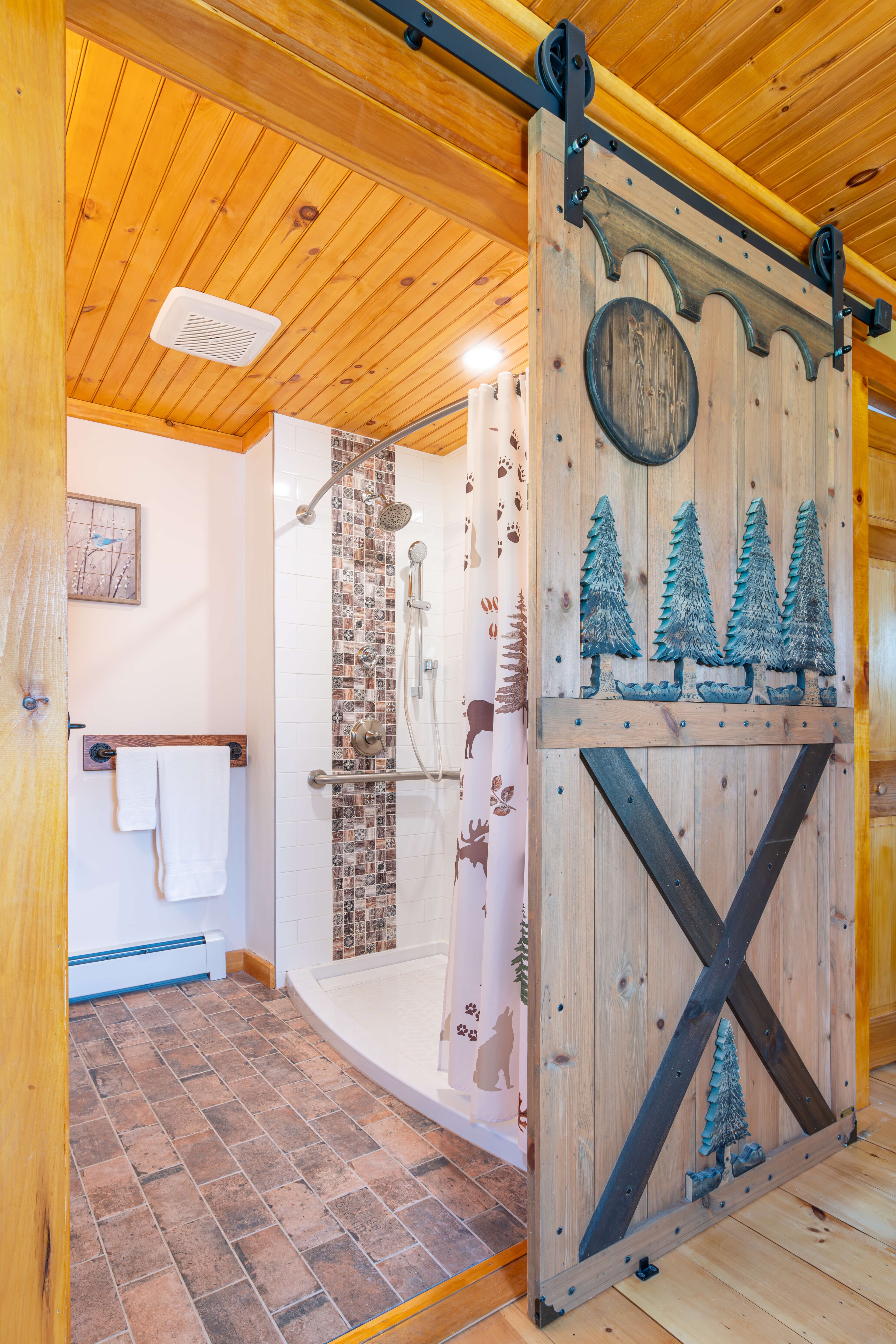 Sliding barn door decorated with pine trees leading into ADA bathroom with walk in shower and handrail.