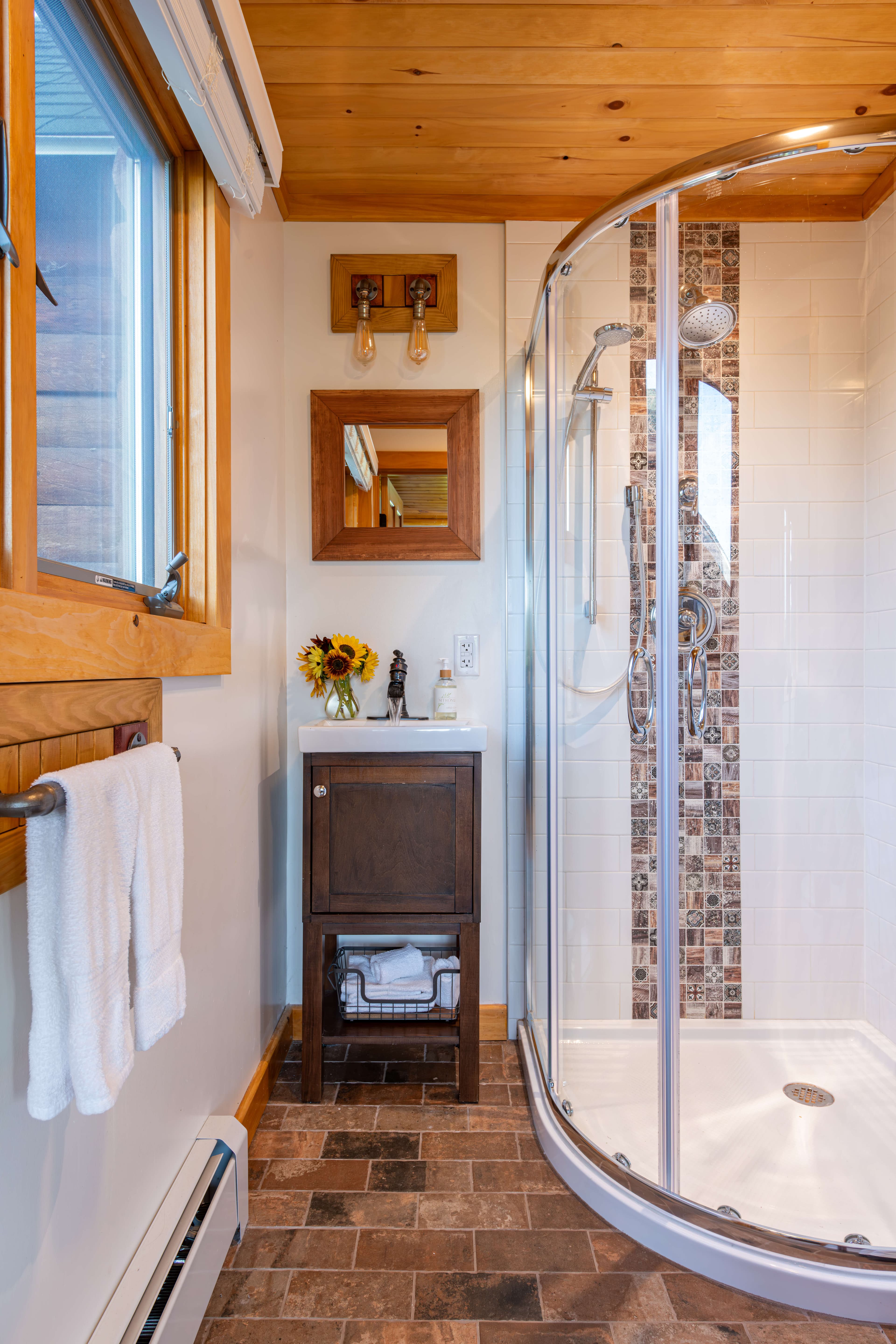 Glass shower on brick floor and wooden mirror over sink