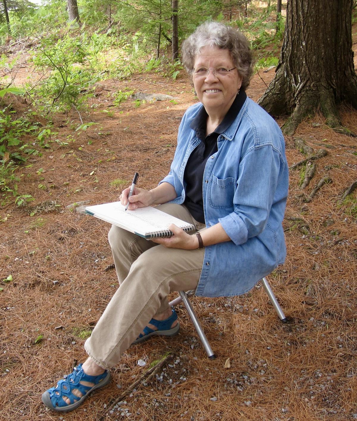 A woman in a blue shirt sitting in the woods with a sketchbook