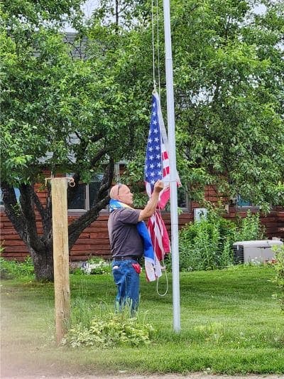 A man in jeans and t-shirt with sunglasses on his head, raising the flag on a pole next to a hitching post.