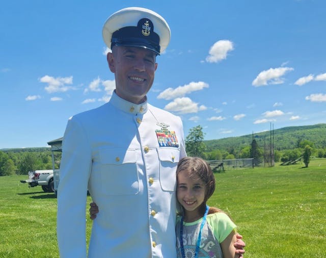 A soldier in a white uniform being hugged by a little girl standing in grass under clear sky