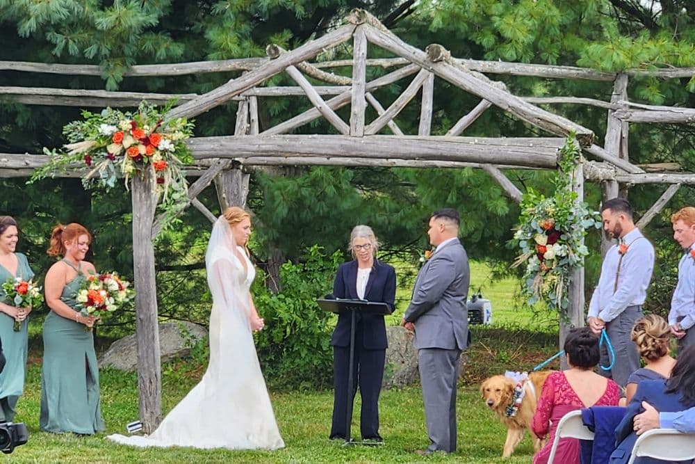 A bride and groom and officiant standing outdoors by a rustic wooden arch in front of large trees