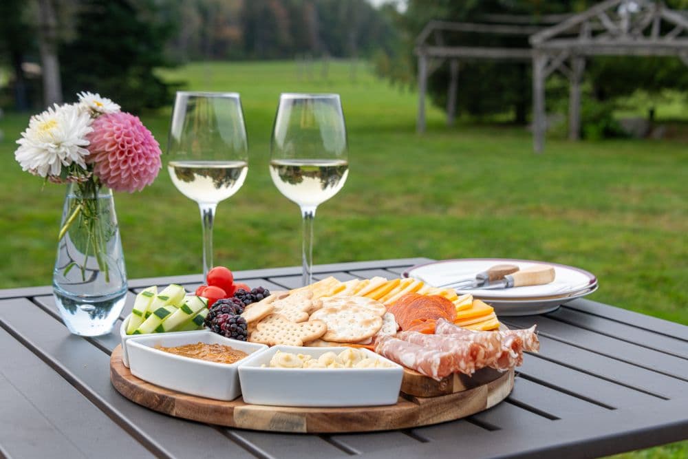 Charcuterie tray, two glasses of white wine, and vase of flowers on a small table outdoors.