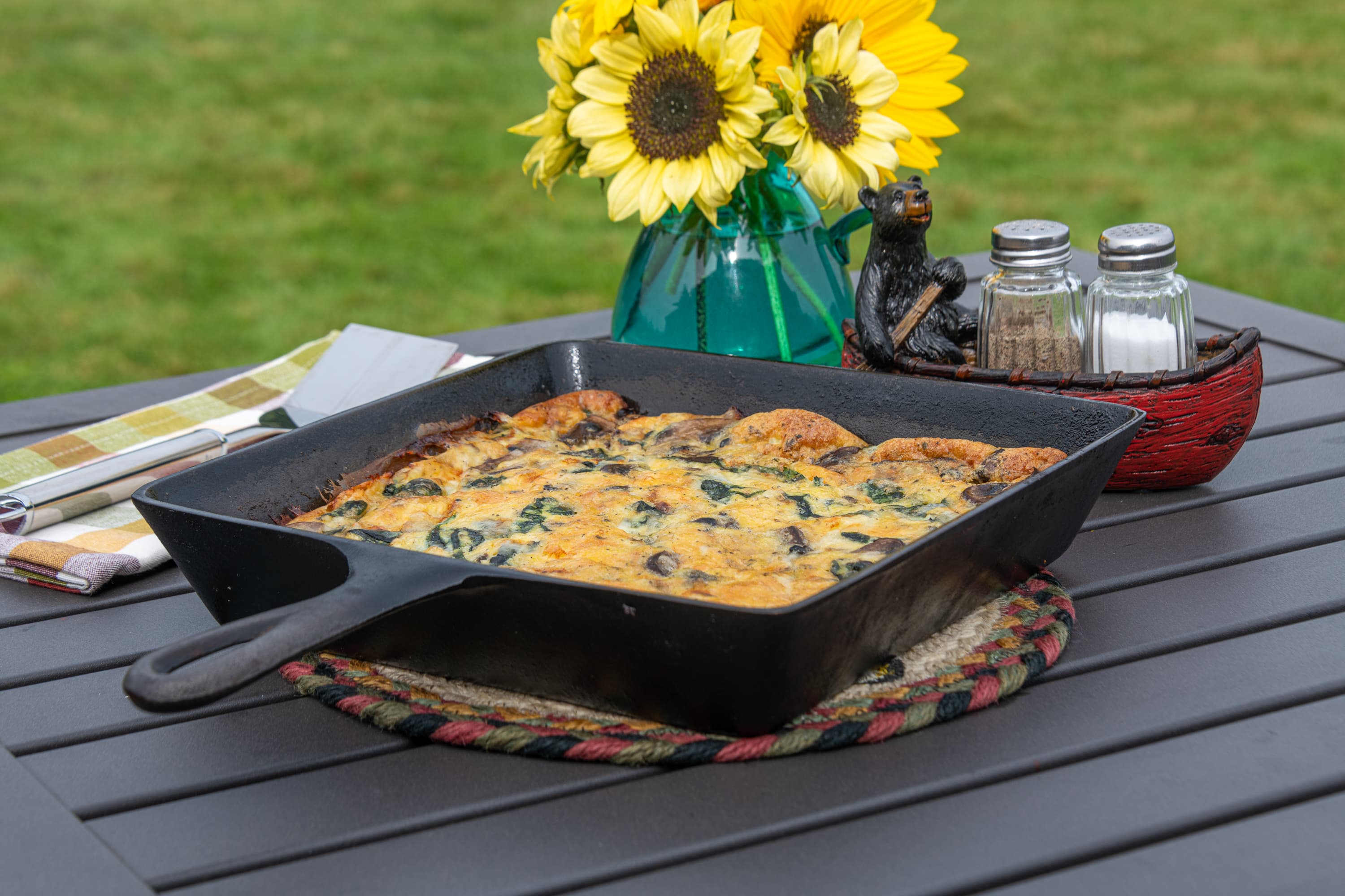 Quiche in a square pan, on a picnic table with sunflowers in a vase, with salt and pepper serving containers