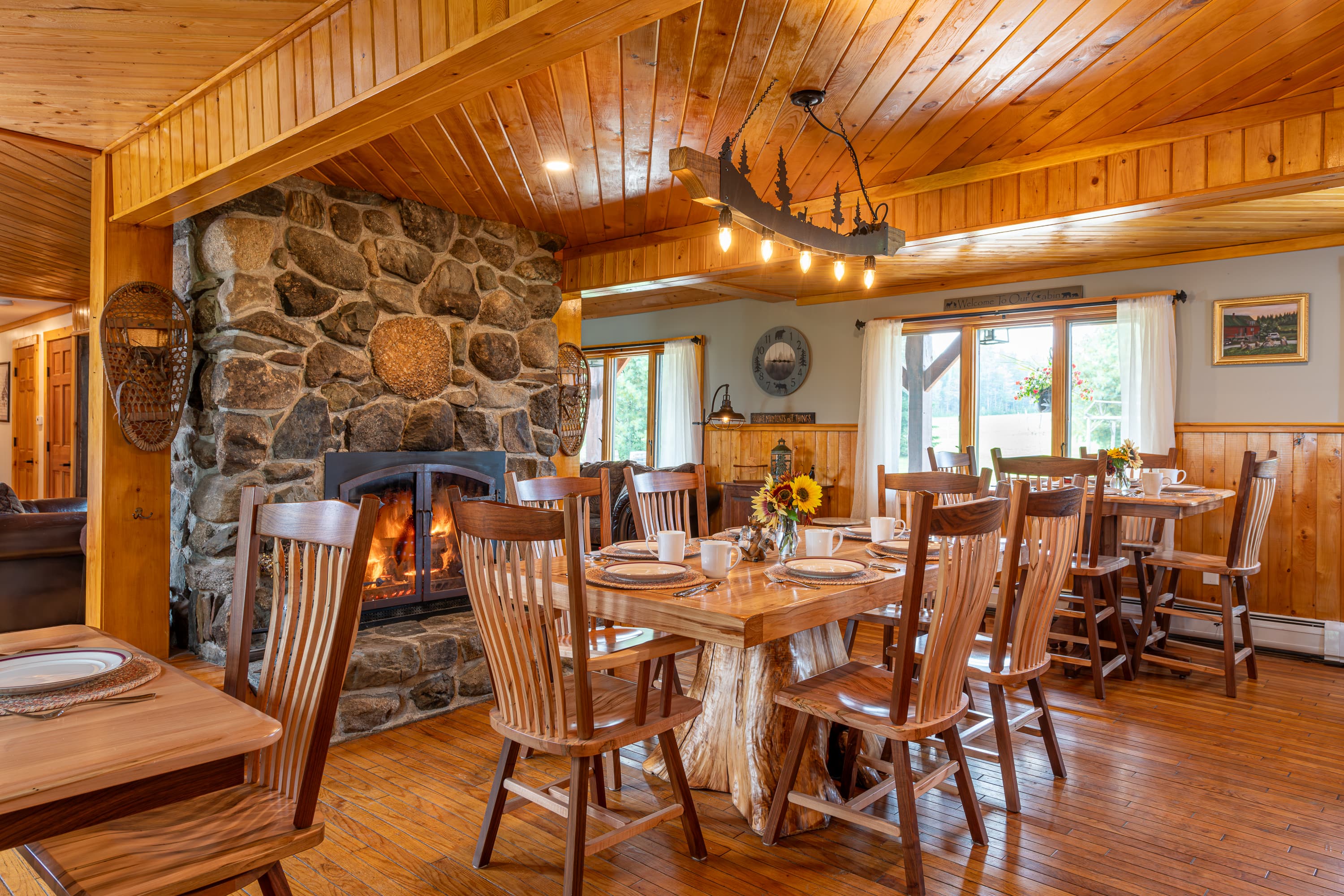 Large wood kitchen table and chair with a blazing stone fireplace and rustic overhead lighting