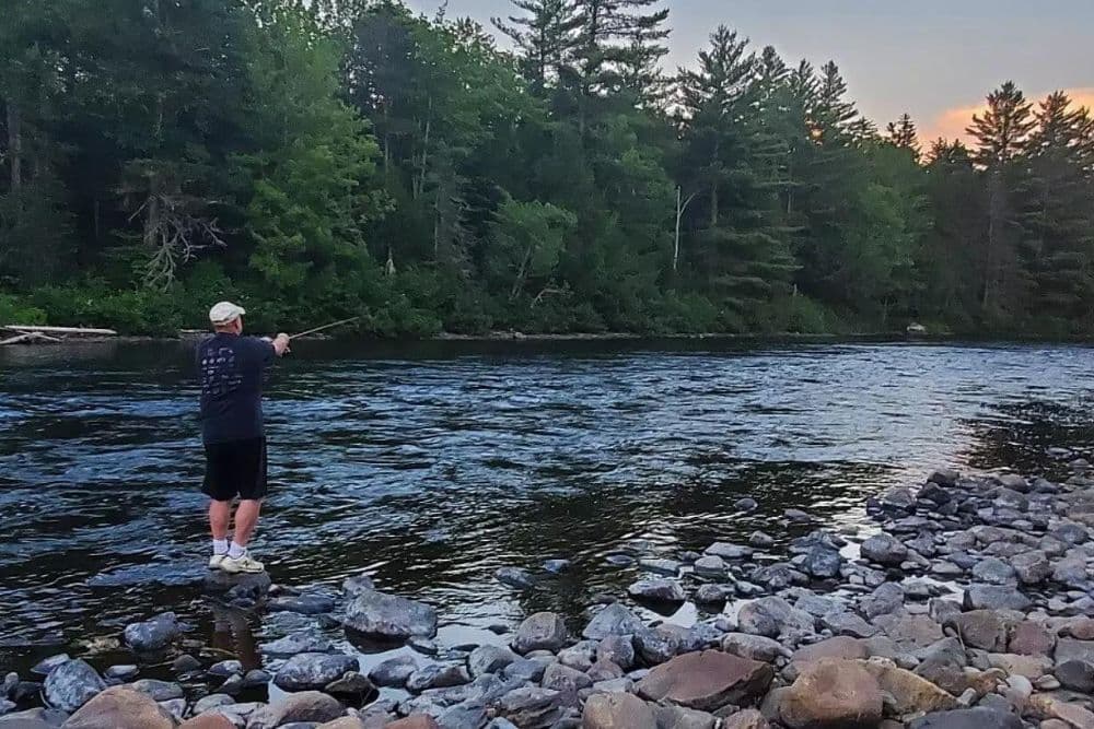 A man standing fishing while standing at the edge of a river with a rocky shore