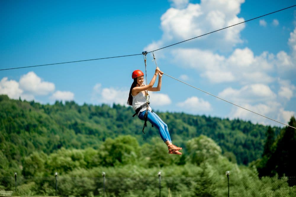 A woman in blue pants, white top and red helmet swinging down a zip line surrounded by trees