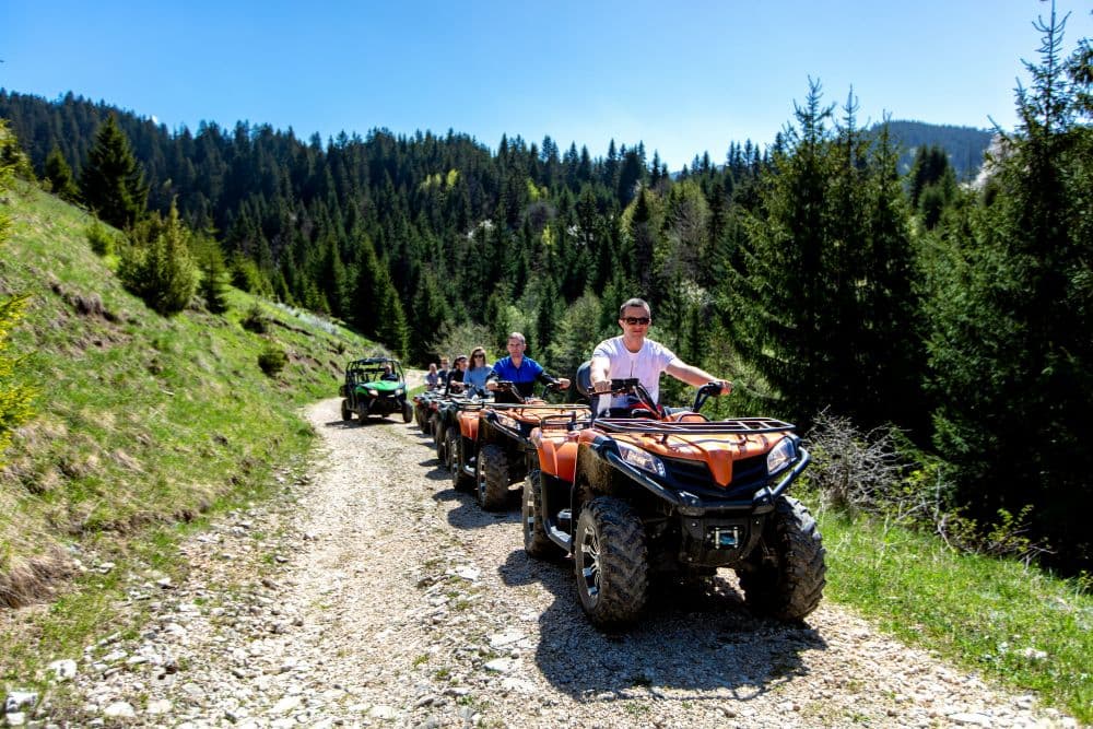 A group of people, each riding ATV machines in a row on a gravel path in the mountains