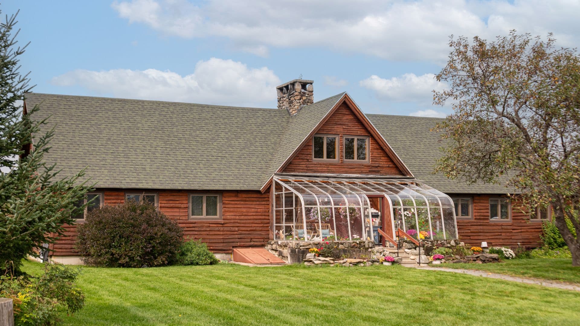 Front view of a large log cabin home with a greenhouse feature by the front door, expansive lawn, with lots of trees and bushes