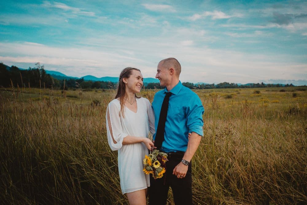 A girl in a white dress holding a bouquet of sunflowers with a guy in a blue shirt and black pants in a large field