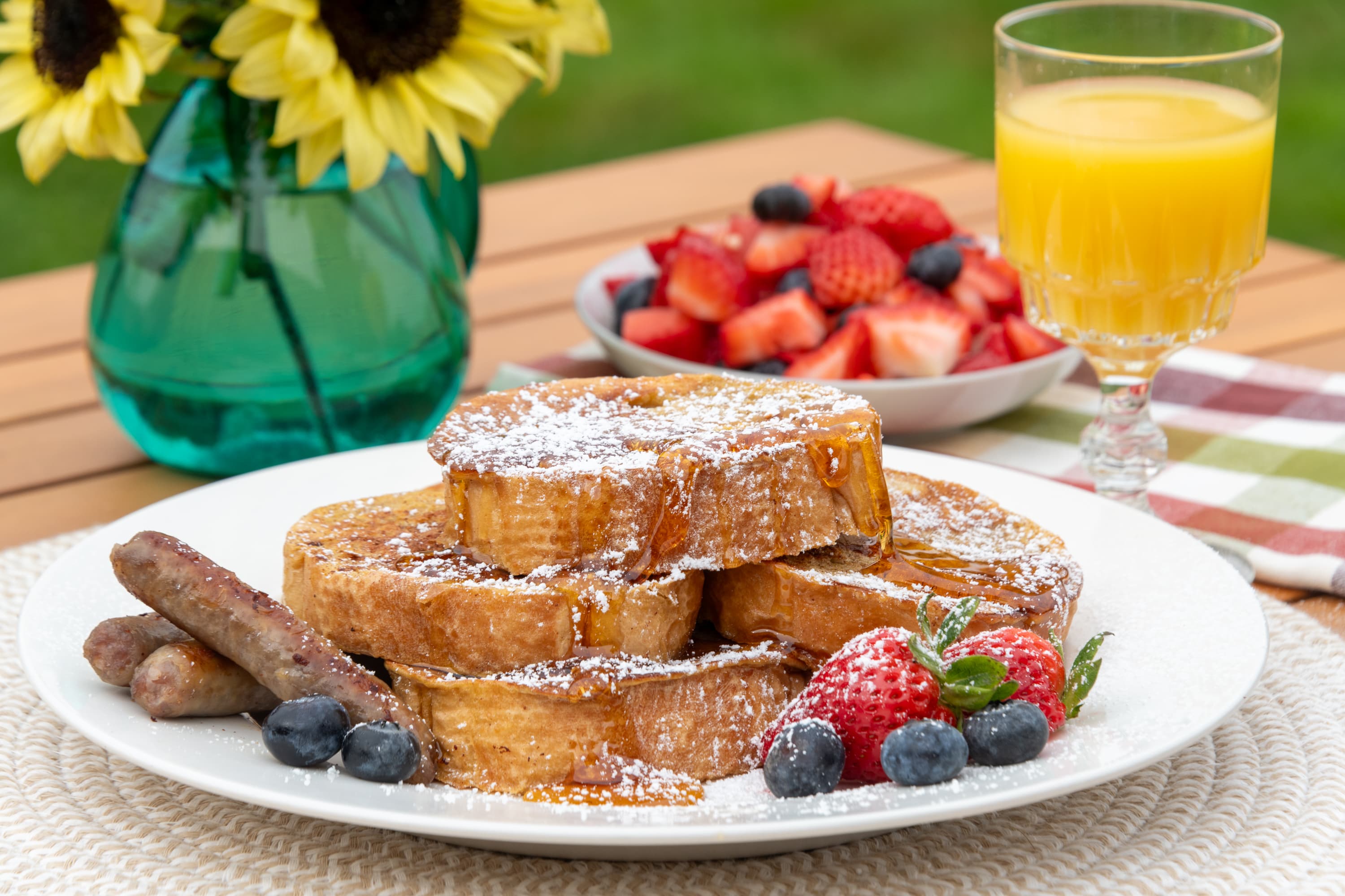 French toast and powdered sugar, glass of orange juice, strawberries, sunflowers in a vase on an outdoor table