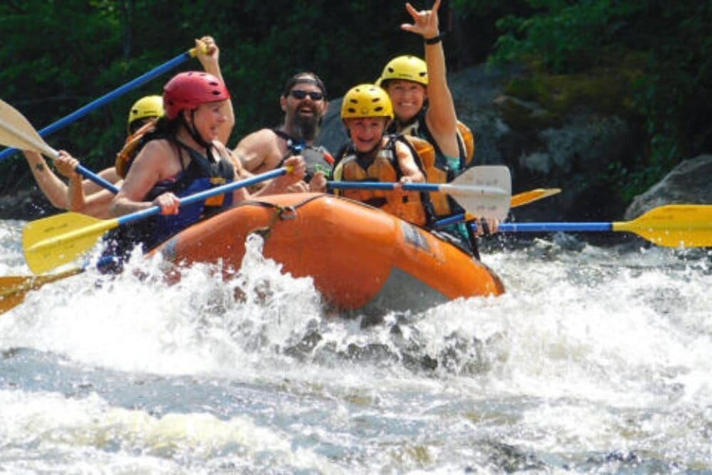 A group of people wearing helmets and holding paddles going down rapids in an orange raft