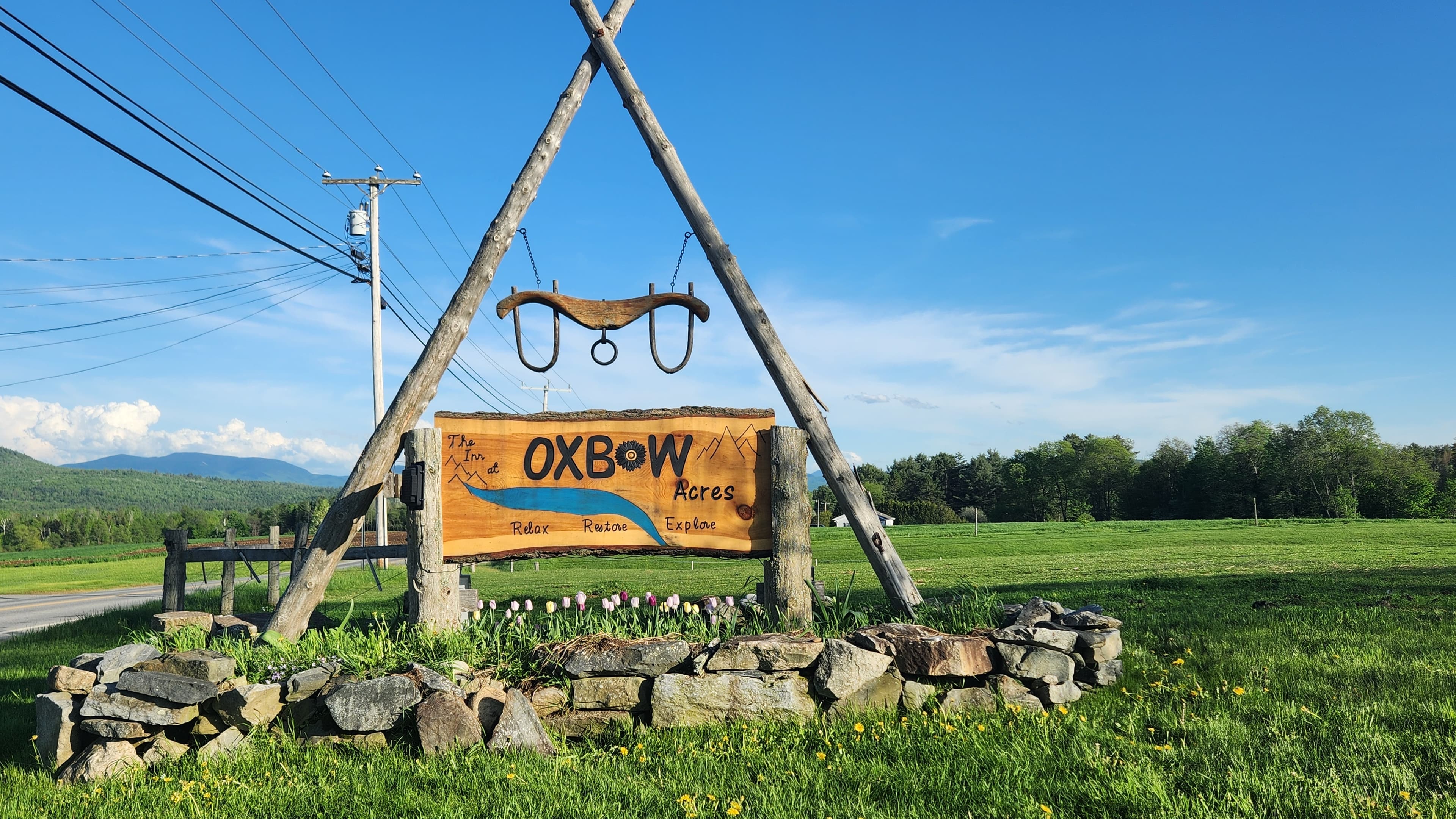 The oxbow acres sign hanging from 2 crossed poles, with a oxbow above them and green grass and mountains in the background