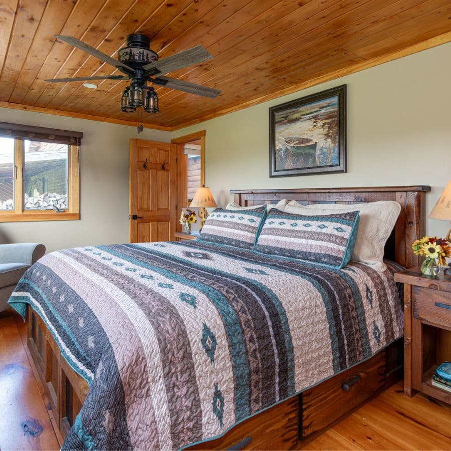 Bedroom with pine headboard, bed with multi color quilt, nightstands with lamps and vases of freshly cut flowers, ceiling fan, door and window