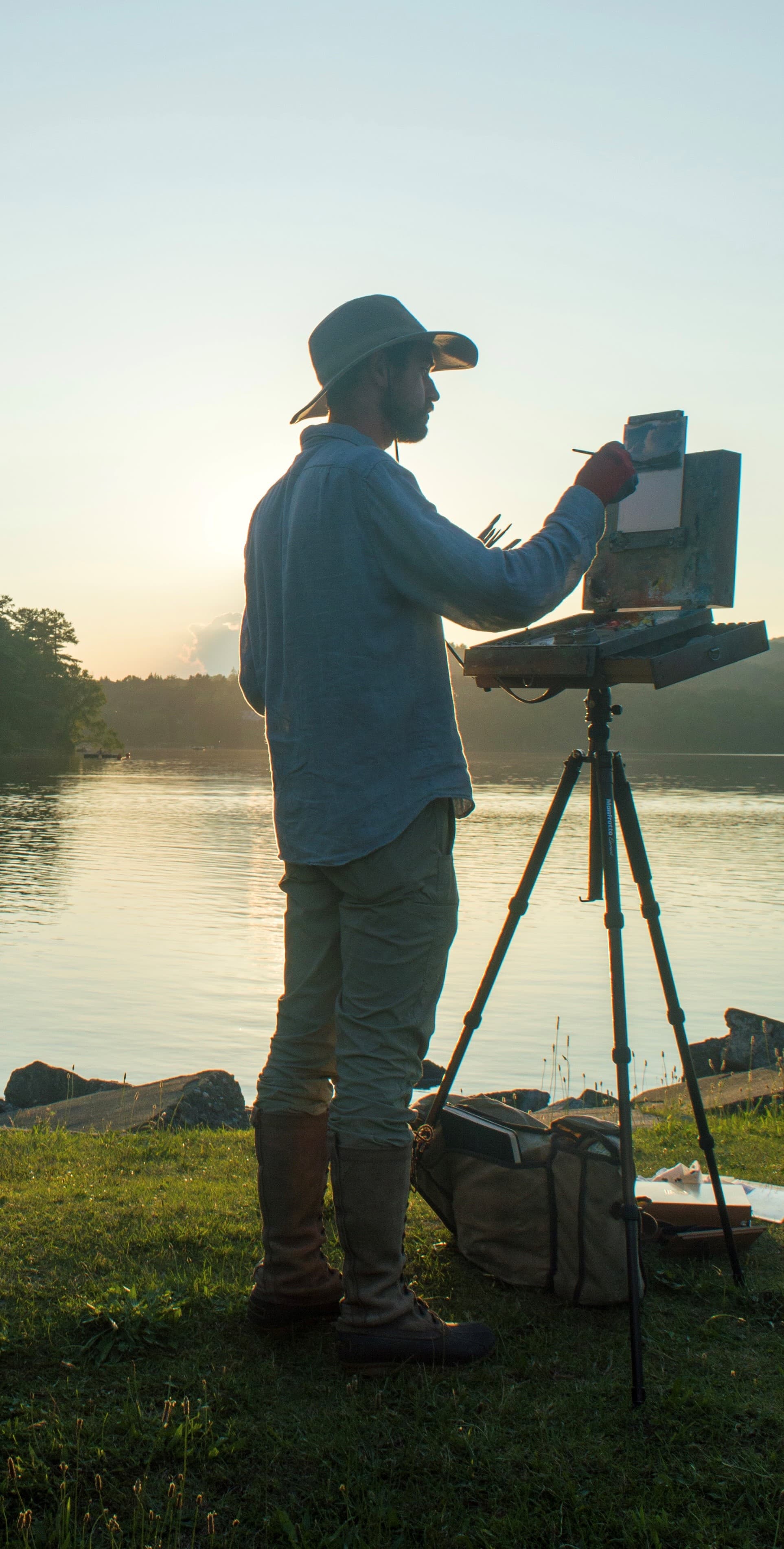 An artist paints at an easel by a serene lake during sunset.