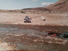 A pickup truck is partially submerged in a fast-flowing river, with a group of people on a vehicle in the background.