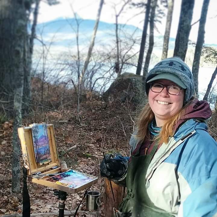 A smiling artist stands outdoors by her easel, surrounded by trees and a scenic view.