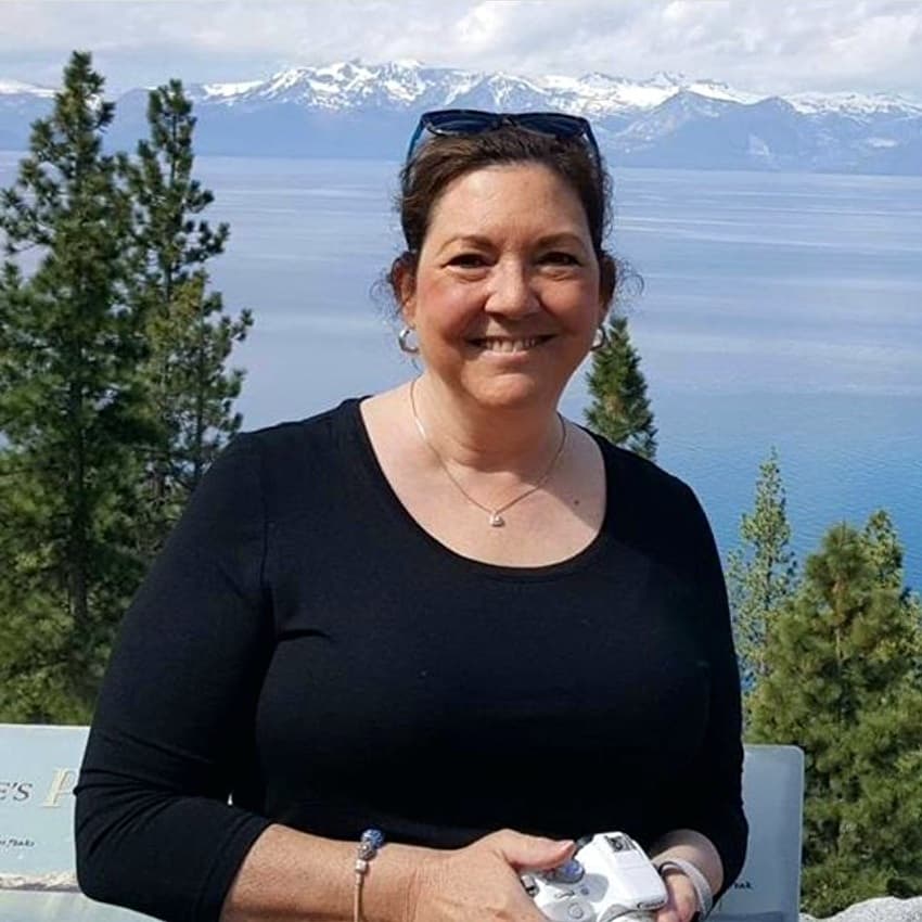 A smiling woman stands outdoors with a scenic lake and mountains in the background, holding a camera.