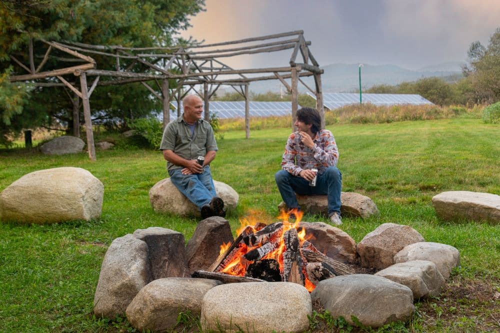 Two people sitting outdoors on large boulders next to a large fire pit.