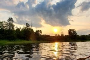 A serene sunset reflecting on a calm river surrounded by lush greenery and clouds.