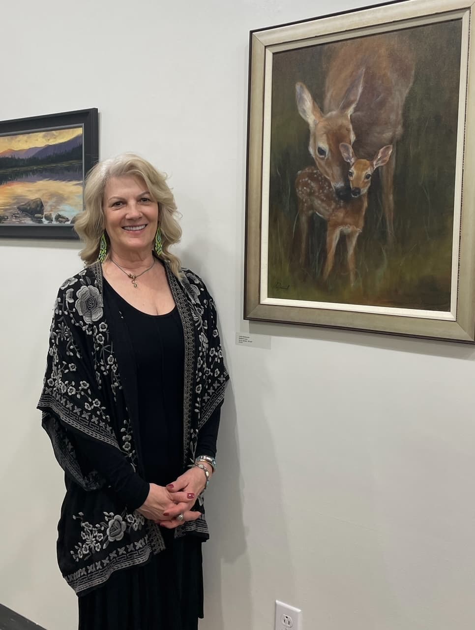A smiling woman in a black outfit stands beside a painting of a deer and fawn in an art gallery.